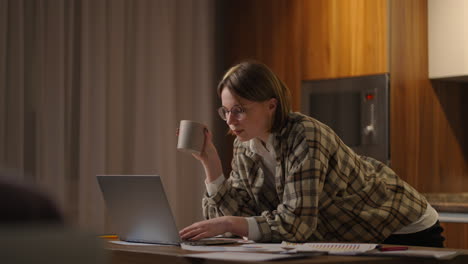A-young-beautiful-business-woman-works-from-home-with-a-cup-of-coffee-and-looks-into-a-laptop-screen-standing-at-the-table.-View-mail-and-drink-coffee.-Remote-work-from-home